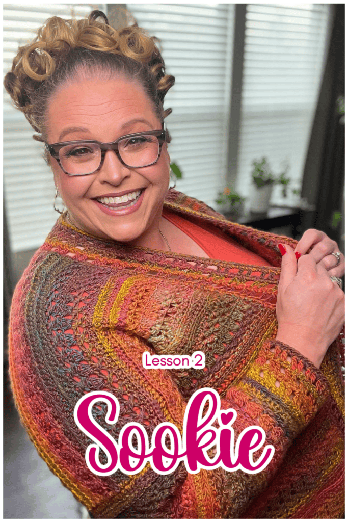 A woman with curly hair and glasses smiles while wearing a colorful, textured shawl, akin to a crochet cardigan. The text "Lesson 2 Sookie" appears on the image. She is standing in a room with large windows in the background, creating an inviting atmosphere for customizing length options. -Marly Bird