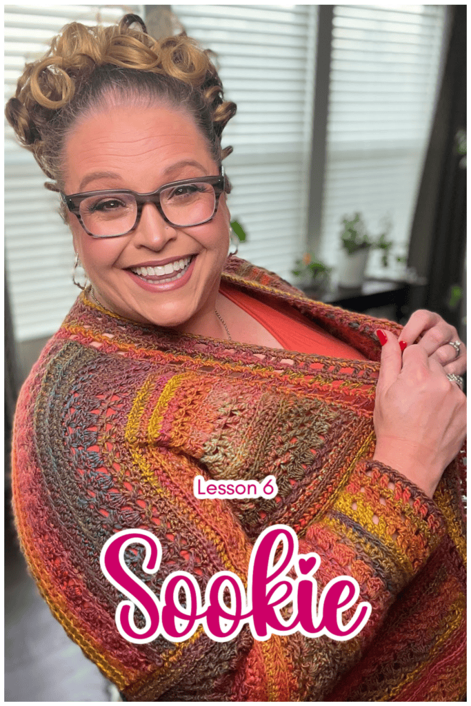 A smiling person wearing glasses and a colorful crocheted shawl over an orange top stands in a bright room. Text on the image reads, "Lesson 6 Sookie." The vibrant scene adds perfect finishing touches to their look. -Marly Bird