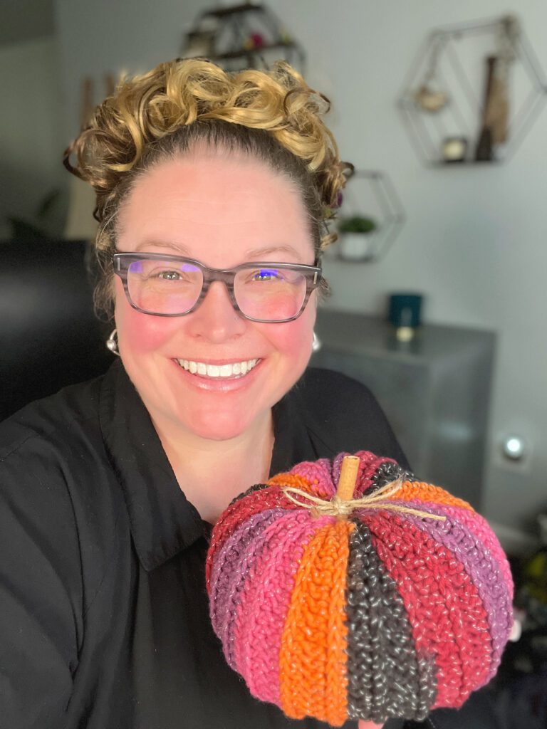 A person with glasses and curled hair, smiling and holding a crocheted pumpkin that features sections of red, orange, yellow, and black. The background includes geometric wall decor and various items on a shelf. -Marly Bird