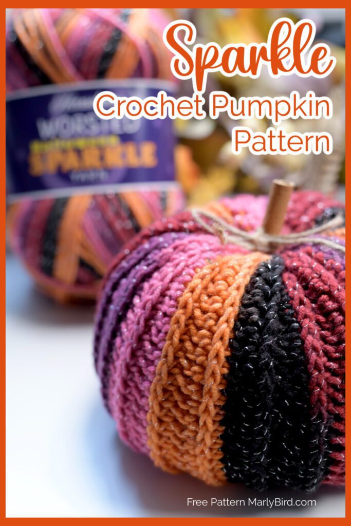 Image showing a crocheted pumpkin made with yarn in alternating stripes of orange, black, and pink, presented on a white surface. In the background, there is a skein of similarly colored yarn. The text on the image reads "Sparkle Crochet Pumpkin Pattern" and "Free Crochet Pumpkin Pattern by Marly Bird. -Marly Bird