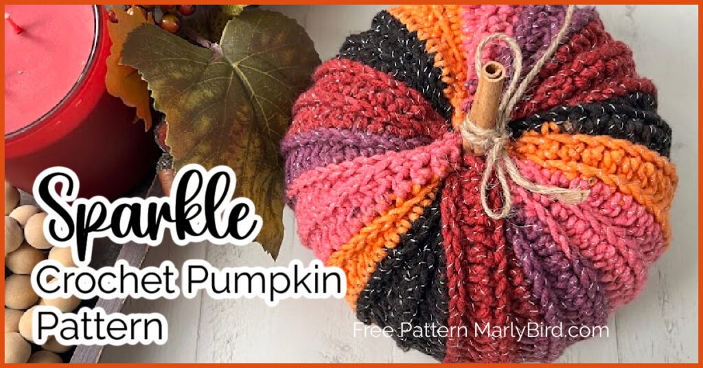 A crocheted pumpkin in vibrant shades of orange, pink, red, purple, and black with a cinnamon stick stem is on display. The pumpkin is accompanied by an orange candle and autumn leaves. Text overlay reads, "Sparkle Crochet Pumpkin Pattern. Free Crochet Pumpkin Pattern MarlyBird.com. -Marly Bird