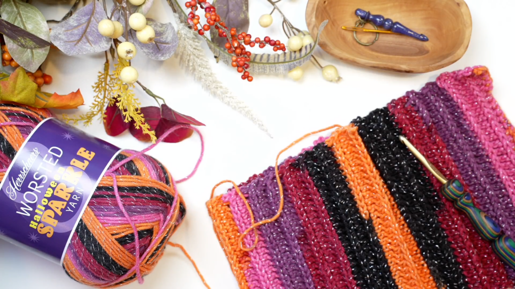 A colorful crochet project with stripes of orange, pink, purple, and black yarn lies partially completed on a white surface. Nearby, a skein of multicolored yarn, autumnal foliage, berries, crochet hooks, and a wooden dish with other crochet tools are arranged neatly next to a crochet pumpkin pattern. -Marly Bird