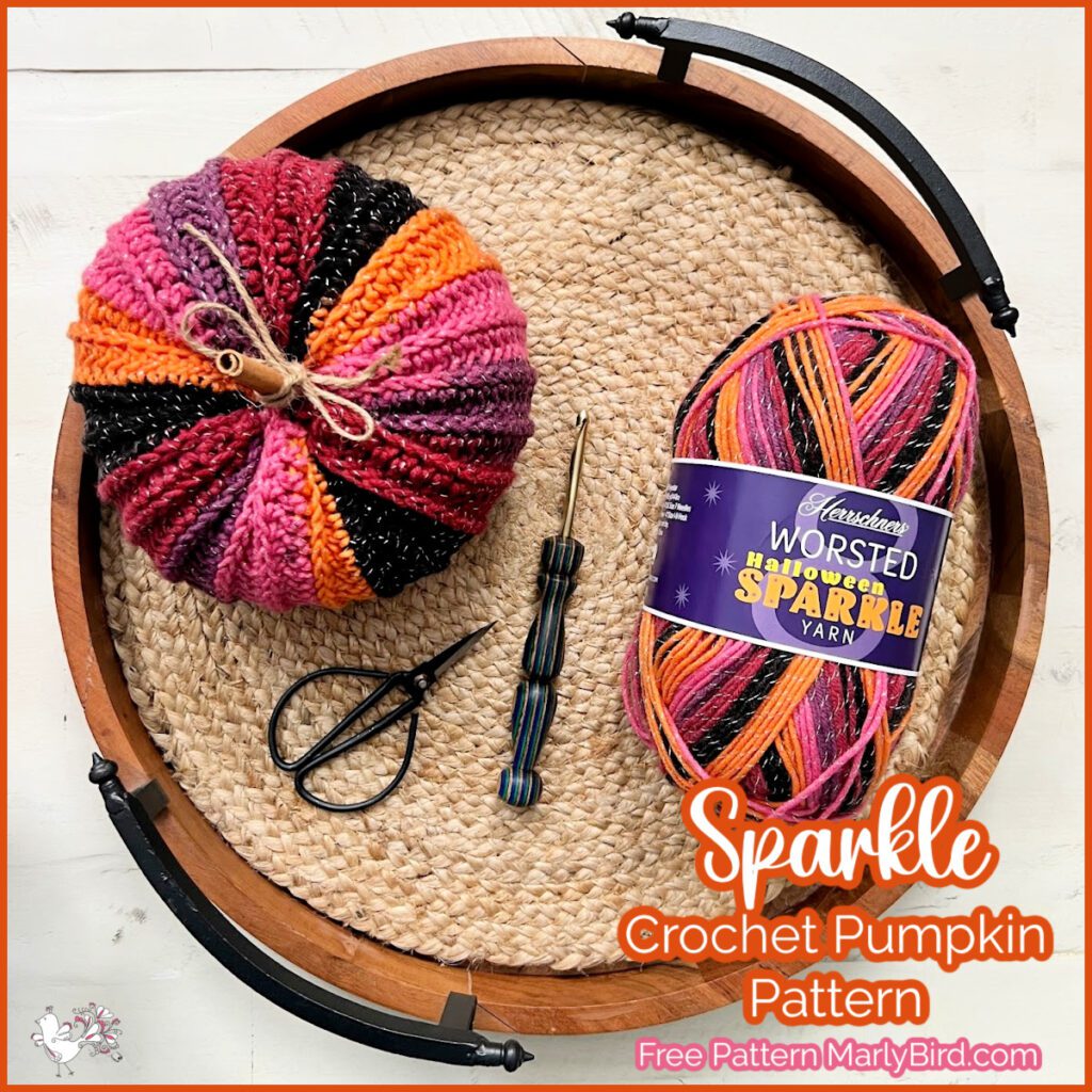 A round woven tray holds a crochet pumpkin in shades of pink, orange, and black, a pair of scissors, a crochet hook, and a skein of variegated yarn labeled "Worsted Show Your Shine Sparkle Yarn." Text on the image reads, "Sparkle Crochet Pumpkin Pattern," showcasing Marly Bird's free crochet pumpkin pattern. -Marly Bird