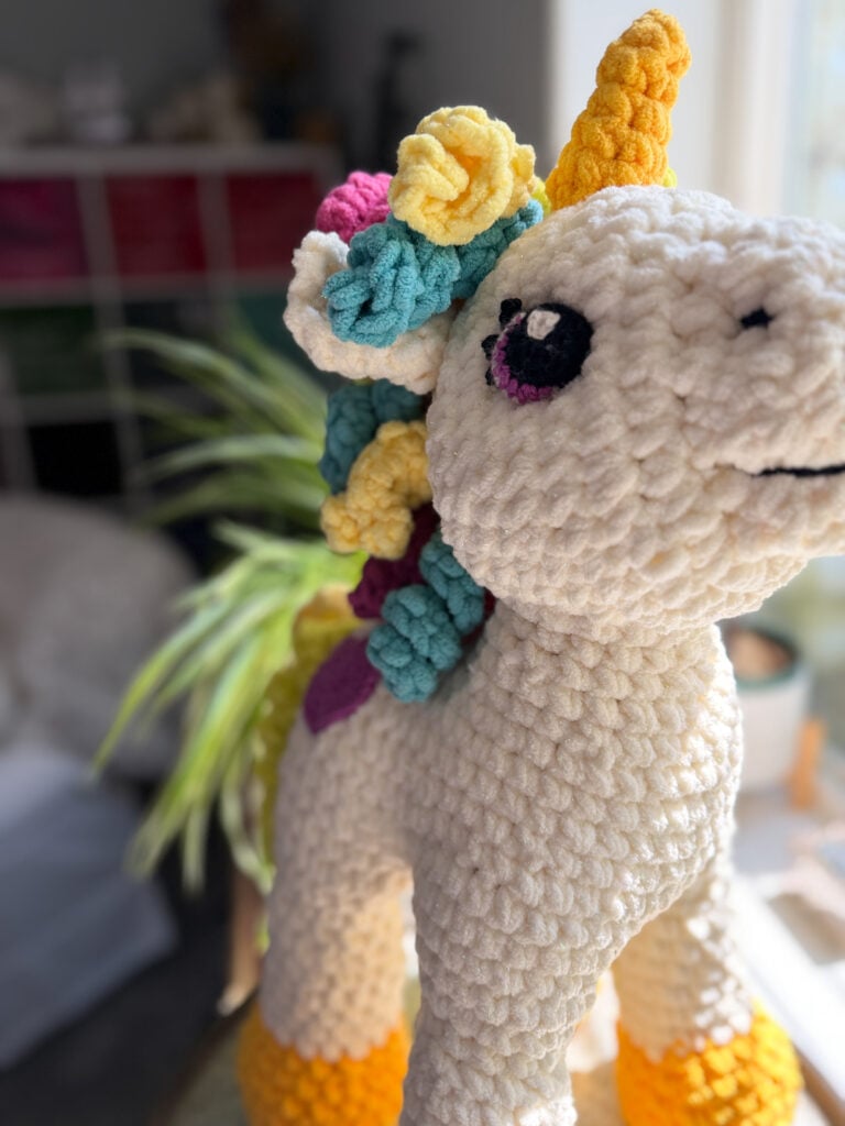Side profile close-up of Sparkle the Crochet Unicorn — cream chenille texture, embroidered eye, yellow spiral horn, and multicolor curly mane, free crochet pattern by Marly Bird