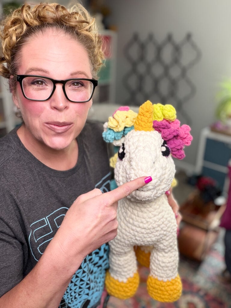 Marly Bird pointing to the face of Sparkle the Crochet Unicorn — showing embroidered eye, gold horn, and rainbow curly mane on the free stuffie pattern