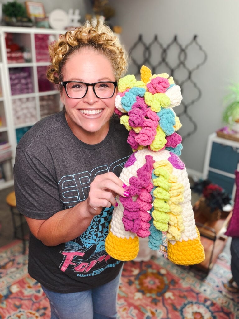 Marly Bird holding Sparkle the Crochet Unicorn upright — 20-inch white amigurumi stuffie with rainbow curly mane, gold horn, and yellow hooves