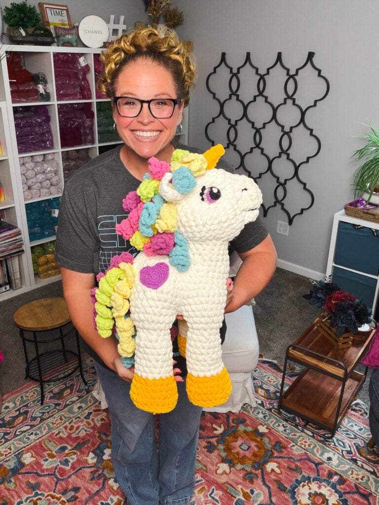 Marly Bird holding Sparkle the Crochet Unicorn full body — cream amigurumi with rainbow mane, purple heart, yellow horn and hooves, 20 inches tall