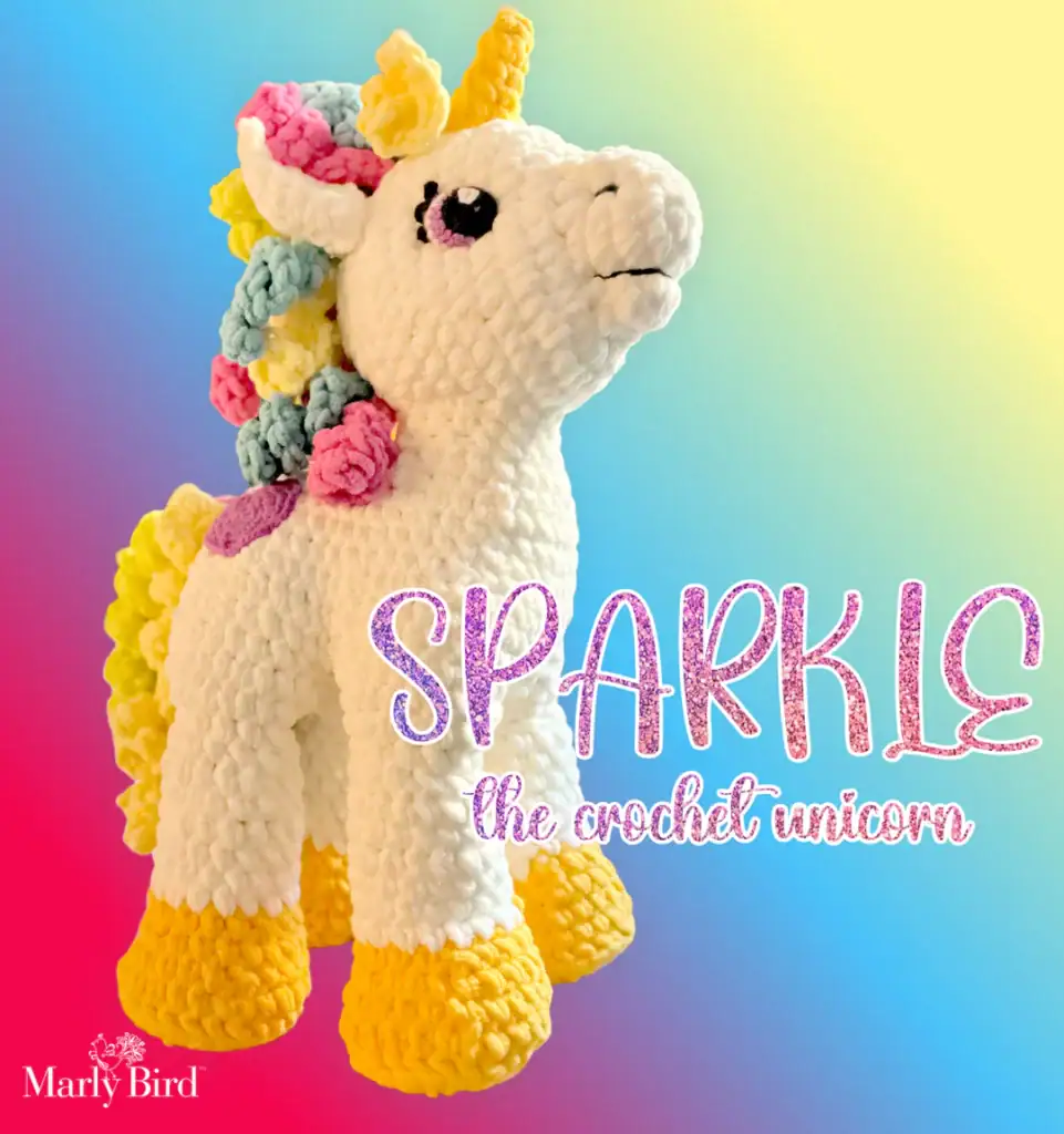 A crocheted unicorn plush with a white body, yellow hooves and horn, and a colorful pastel mane and tail stands in front of a rainbow gradient background. Text reads "SPARKLE the crochet unicorn—find free knitting and crochet patterns. -Marly Bird