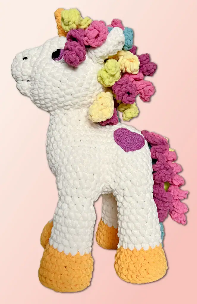 A handmade crocheted unicorn plush toy with a white body, yellow hooves, purple heart detail, and a multicolored mane and tail in pink, yellow, and purple—perfect for fans of free knitting and crochet patterns. Set against a pink gradient background. -Marly Bird