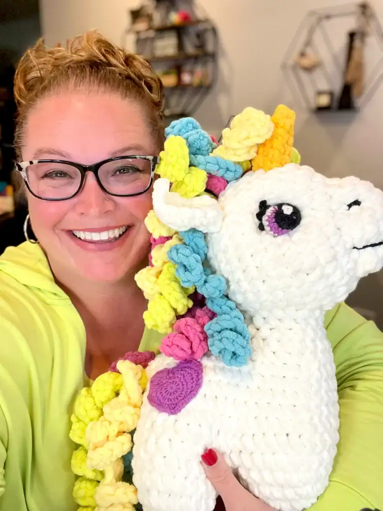 A smiling woman with glasses and a yellow top holds a large, crocheted unicorn with a colorful mane and yellow horn. The softly blurred background shows shelves and decor, hinting at her love for free knitting and crochet patterns. -Marly Bird