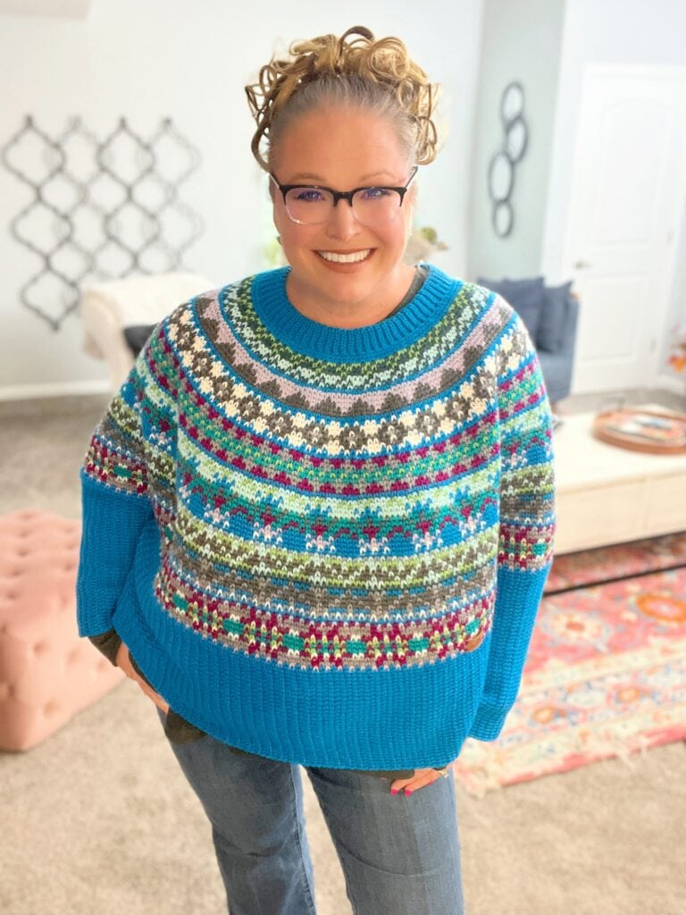 A smiling woman with curly hair and glasses wears a bright, multicolored patterned sweater and blue jeans, standing in a cozy, well-lit living room with modern decor and a patterned rug. -Marly Bird