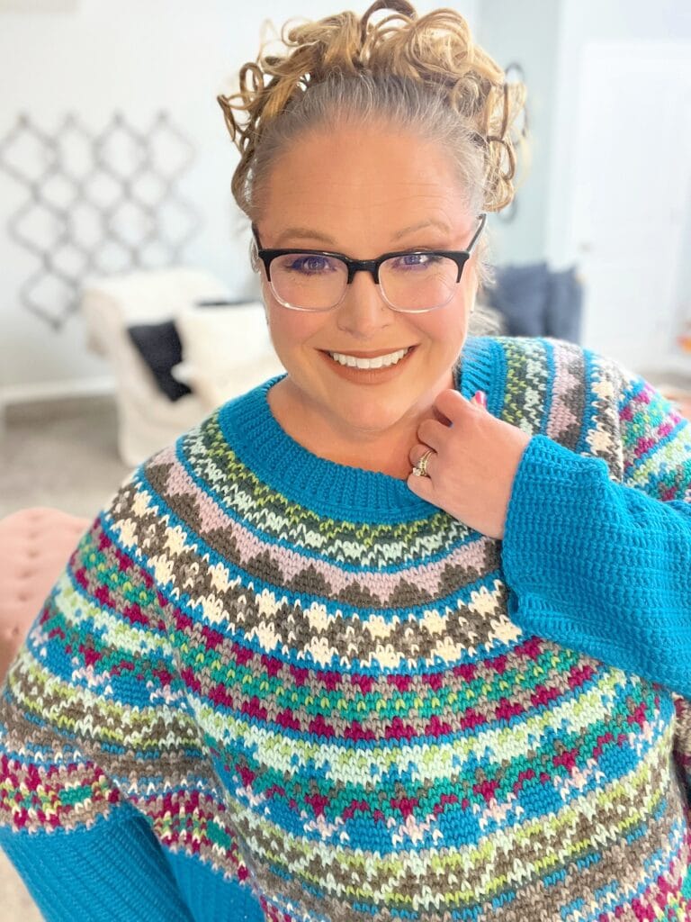 Smiling woman with curly hair, glasses, and a colorful patterned sweater poses indoors. She stands in a bright room with neutral decor, looking at the camera. -Marly Bird