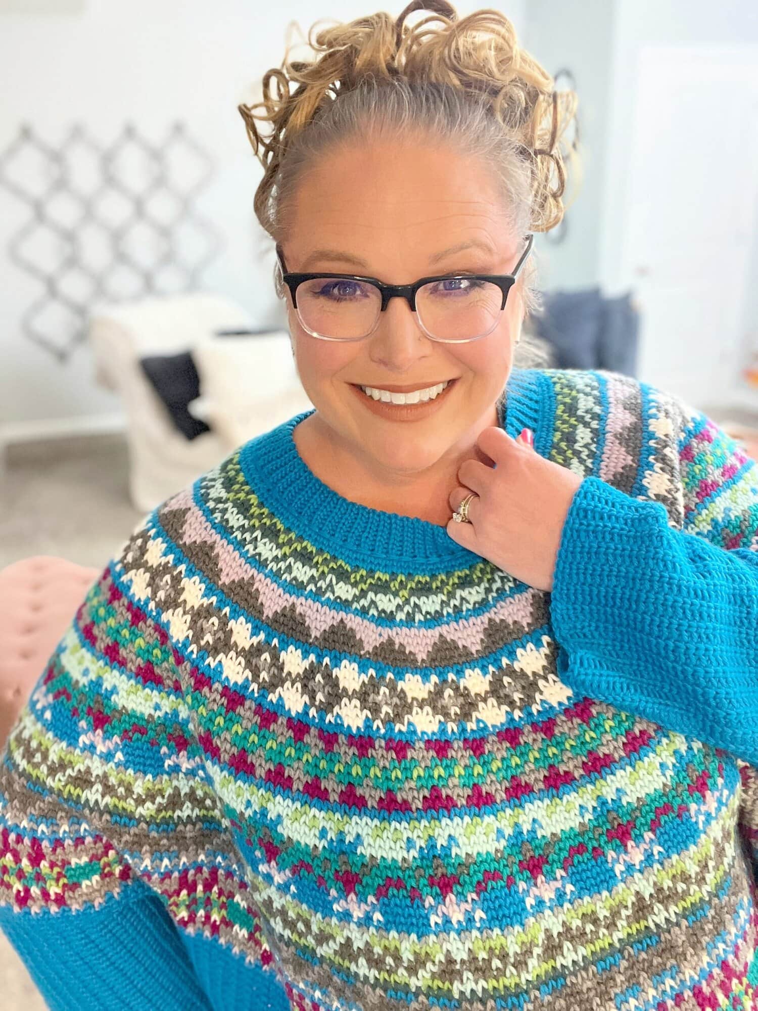 Smiling woman with curly hair, glasses, and a colorful patterned sweater poses indoors. She stands in a bright room with neutral decor, looking at the camera. -Marly Bird