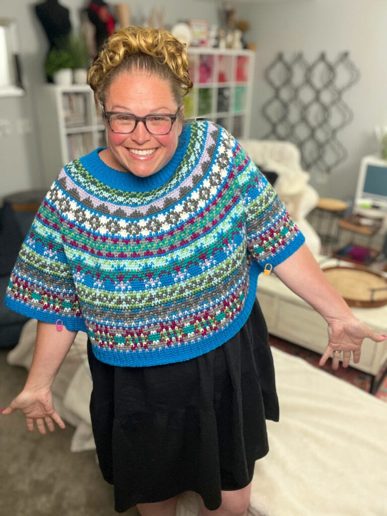 A smiling person with glasses and curly hair wears a colorful, patterned knit sweater over a black dress, standing in a cozy living room filled with yarn and decor—perfect inspiration for your next crochet swoncho or prism make-along project. -Marly Bird