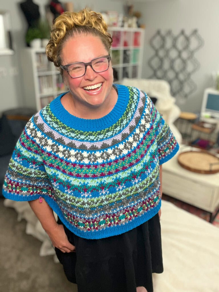 A smiling person with curly hair and glasses wears a colorful Crochet Swoncho over a black dress, standing in a cozy room with shelves, books, and yarn art—perfect inspiration for the Prism Make-Along 2025. -Marly Bird