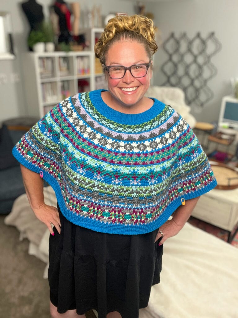 A smiling woman with glasses and curly hair wears a colorful, patterned Crochet Swoncho over a black dress, standing in a cozy, well-lit room with bookshelves and home decor in the background. -Marly Bird