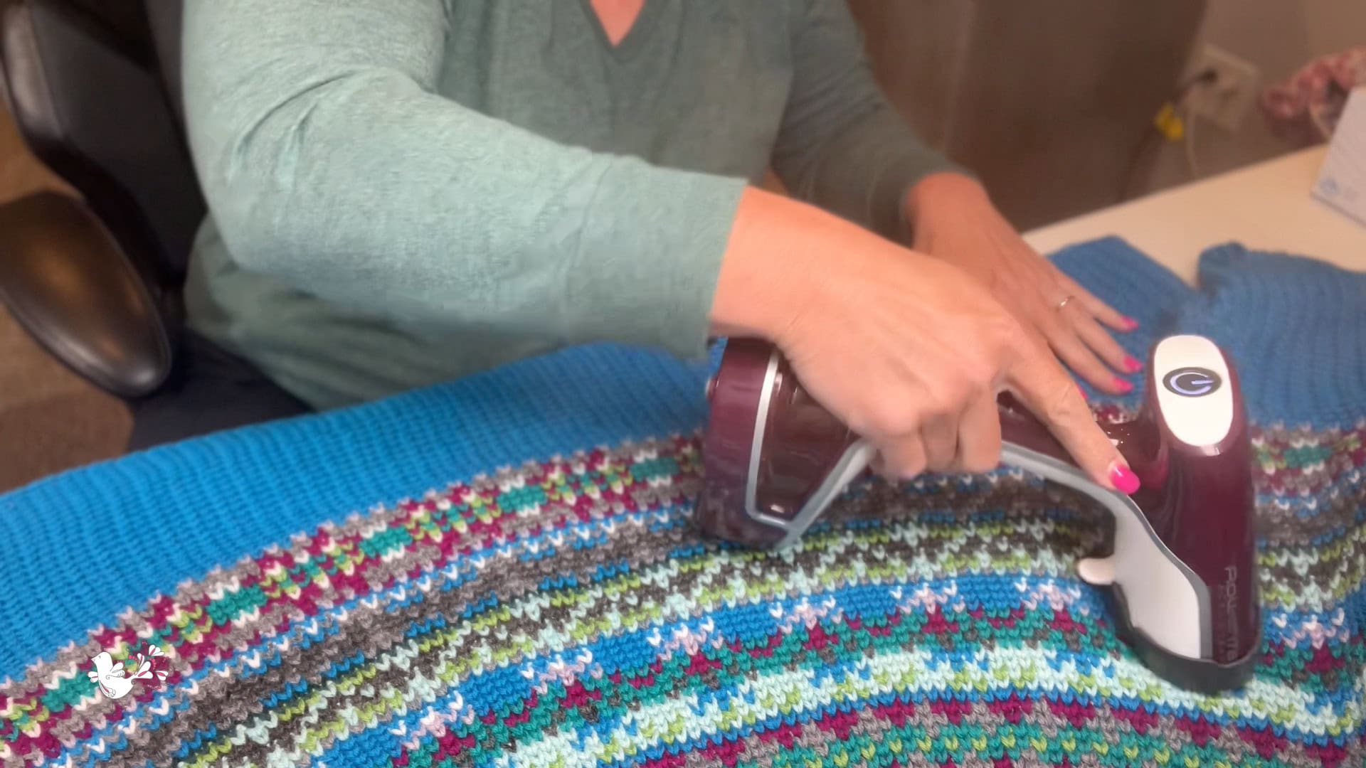 A person in a green long-sleeve shirt uses a handheld fabric shaver to remove fuzz from a colorful, striped blanket made with Superwash Yarn, spread out on a table. -Marly Bird