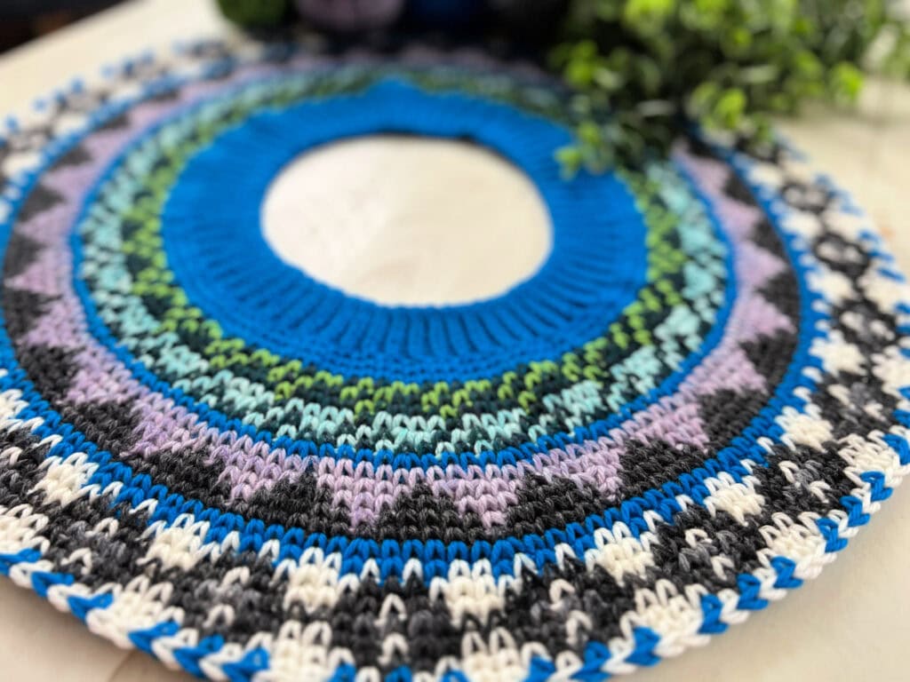 A close-up of a colorful, circular knitted garment with blue, green, purple, black, and white patterns—perfect for a Prism Make-Along—displayed on a light surface next to a leafy green plant. -Marly Bird
