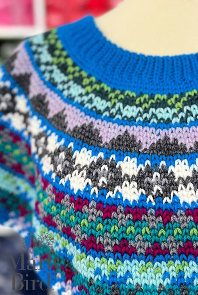 A close-up of a colorful knitted sweater, inspired by Spectrum Stitch Crochet Swoncho Part 4, featuring rows of patterns in blue, purple, green, grey, white, and turquoise yarn with a ribbed blue neckline. -Marly Bird