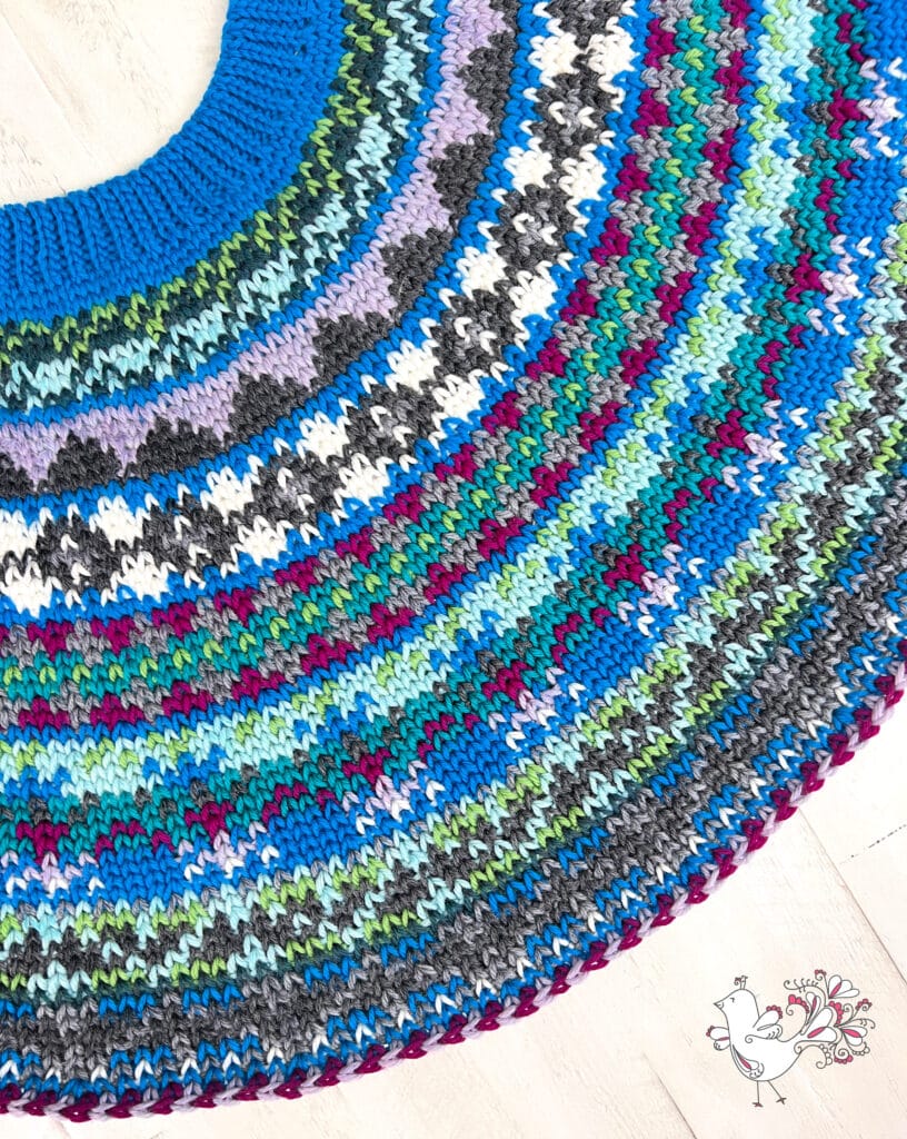 A close-up of a colorful knitted circular pattern from the Prism Make-Along, featuring rows of blue, green, purple, grey, white, and maroon. Detailed geometric motifs and textured stitches shine as a decorative bird illustration appears in the corner. -Marly Bird
