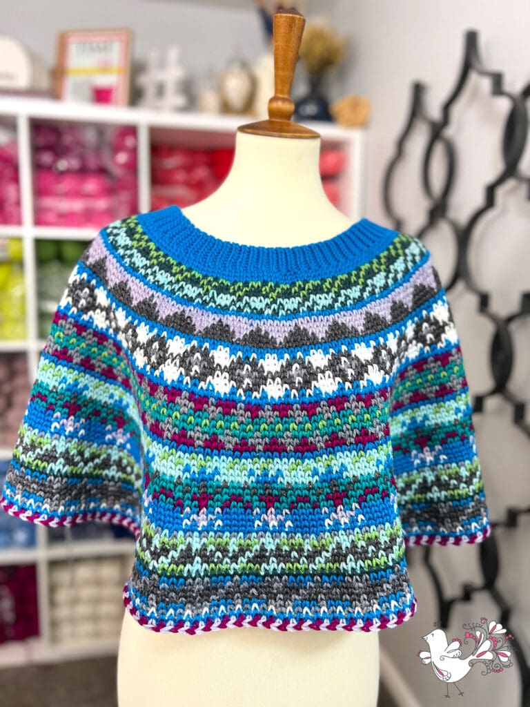 A colorful, short-sleeved knitted sweater with blue, green, red, white, and black patterns is displayed on a dress form. Yarn shelves are visible in the blurred background—perfect for your next Prism Make-Along or Free Pattern project. -Marly Bird