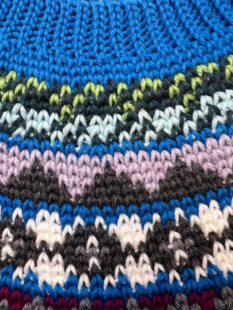 Close-up of colorful knitted fabric featuring zigzag patterns in blue, white, black, purple, green, and turquoise yarns. The textures and stitches are clearly visible—perfect inspiration for your next Spectrum Stitch Crochet project. -Marly Bird