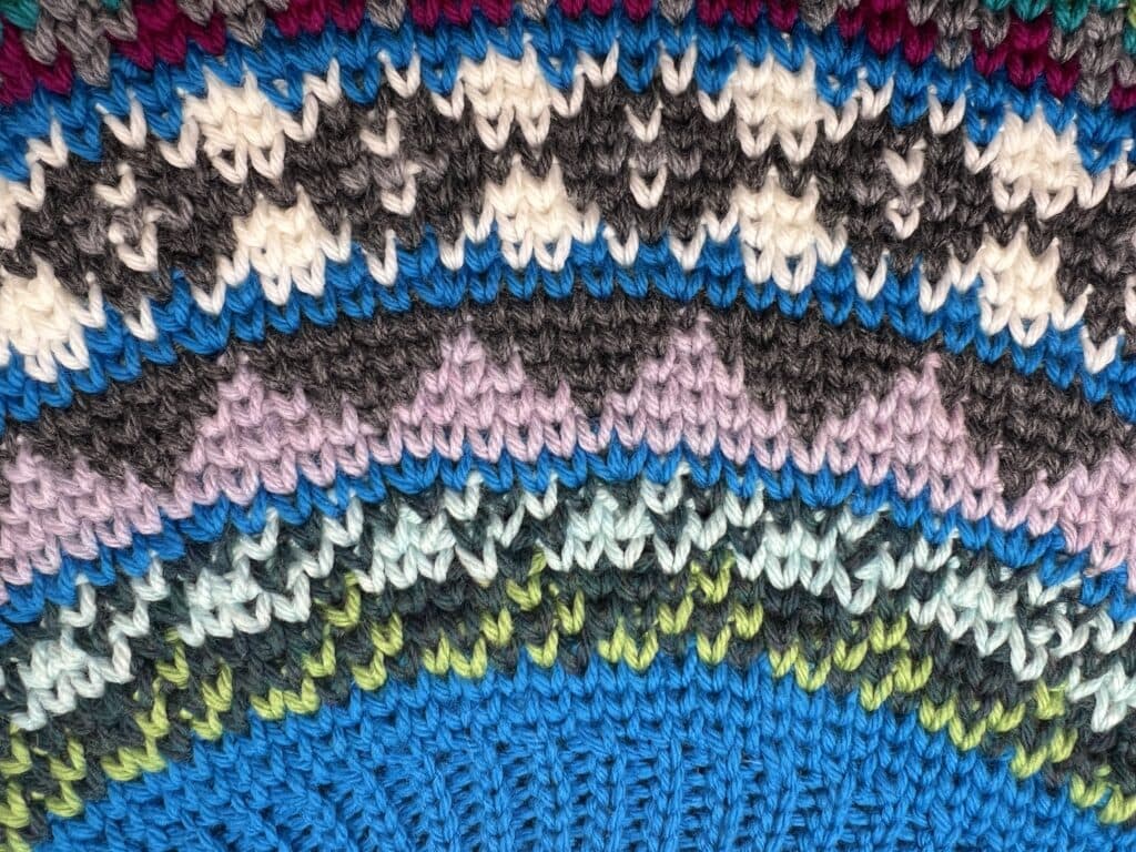 Close-up of colorful knitted fabric with repeating zigzag and wavy patterns in blue, green, purple, black, white, and maroon tones. The stitching creates a textured semicircular design—perfect inspiration for your next Crochet Swoncho or Free Pattern project. -Marly Bird