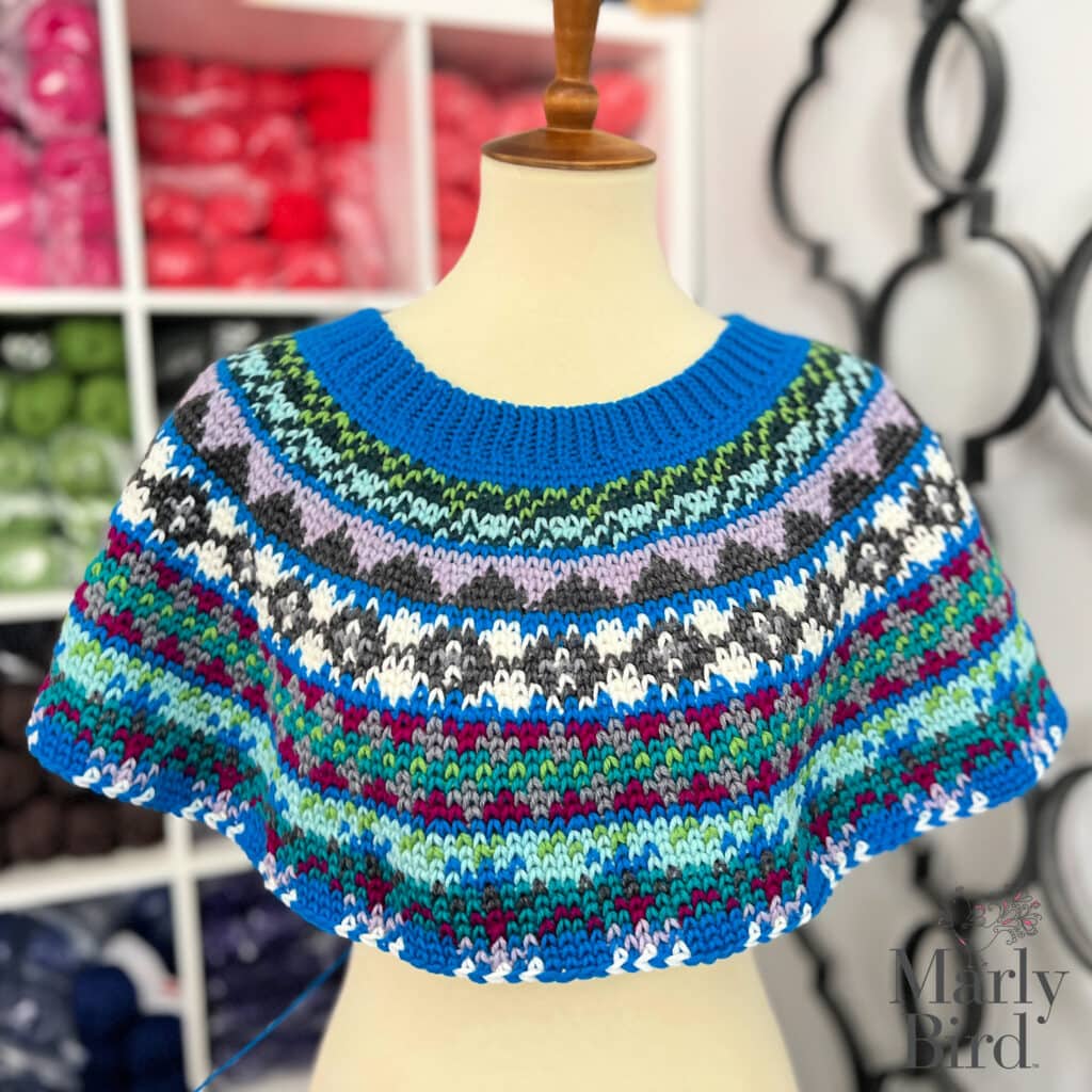 A mannequin displays a colorful, hand-knitted yoke with bold stripes and intricate patterns in blue, green, red, black, purple, and white. Yarn skeins are visible on shelves—perfect inspiration for your Prism Make-Along 2025 project!. -Marly Bird