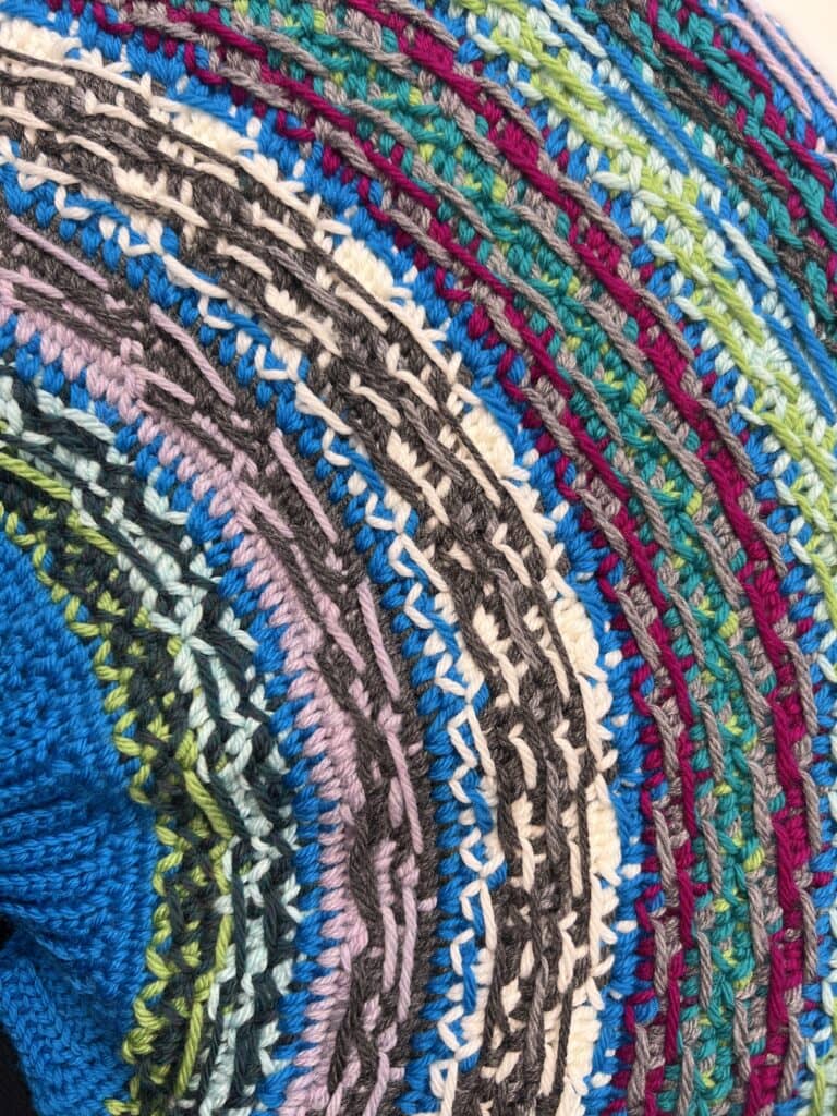 Close-up of colorful, textured knitting with alternating rows of blue, purple, green, white, gray, and burgundy yarn—forming curved stripes in a circular pattern inspired by the vibrant Spectrum Stitch Crochet style. -Marly Bird