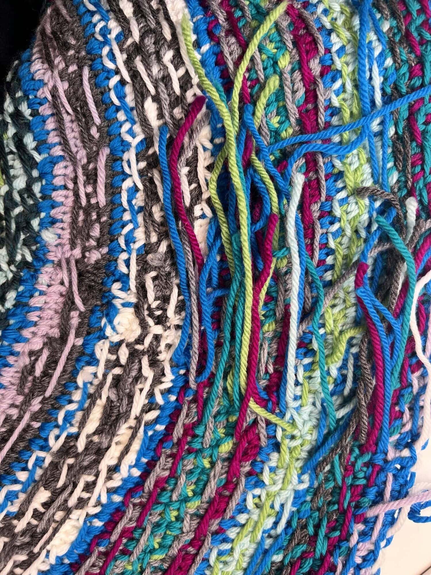 Close-up of colorful yarn strands and crochet stitches in blue, green, purple, white, and black on a partially finished Crochet Swoncho from the Prism Make-Along 2025, with loose yarn ends visible. -Marly Bird