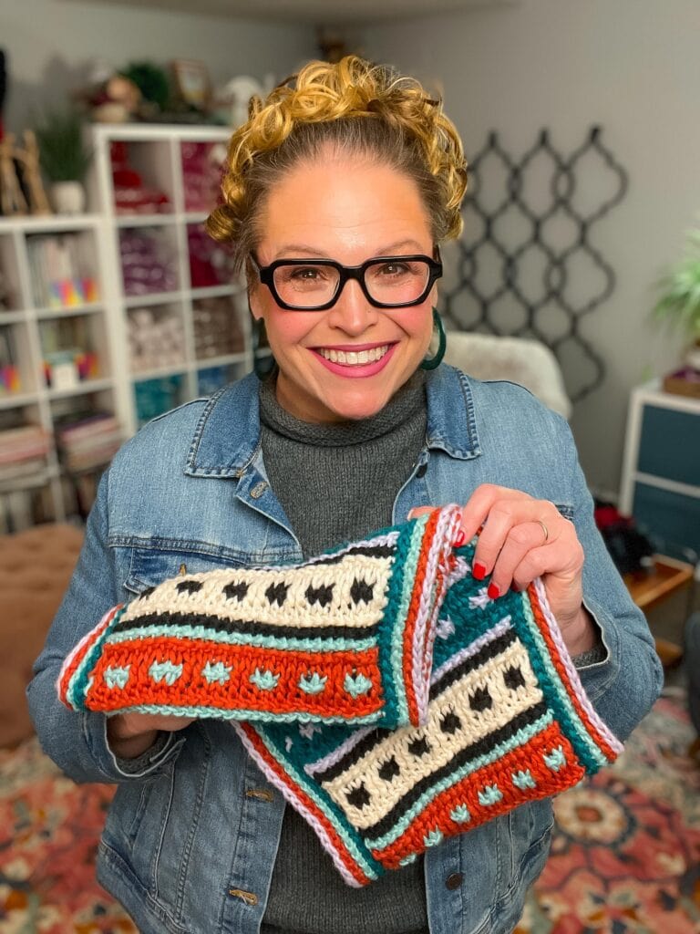 A smiling woman with glasses and curly hair, wearing a denim jacket, holds up a colorful Tunisian Crochet piece with orange, teal, black, and white patterns in a cozy, yarn-filled room. -Marly Bird