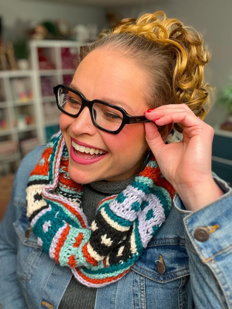 A smiling woman with curly blonde hair wears black glasses, a colorful Tunisian crochet cowl, and a denim jacket. She adjusts her glasses and laughs in a cozy, indoor space—perfect inspiration for your next free pattern!. -Marly Bird