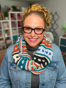 Woman models a vibrant crochet scarf with visible textured stitches; yarn and craft books fill shelves behind her.