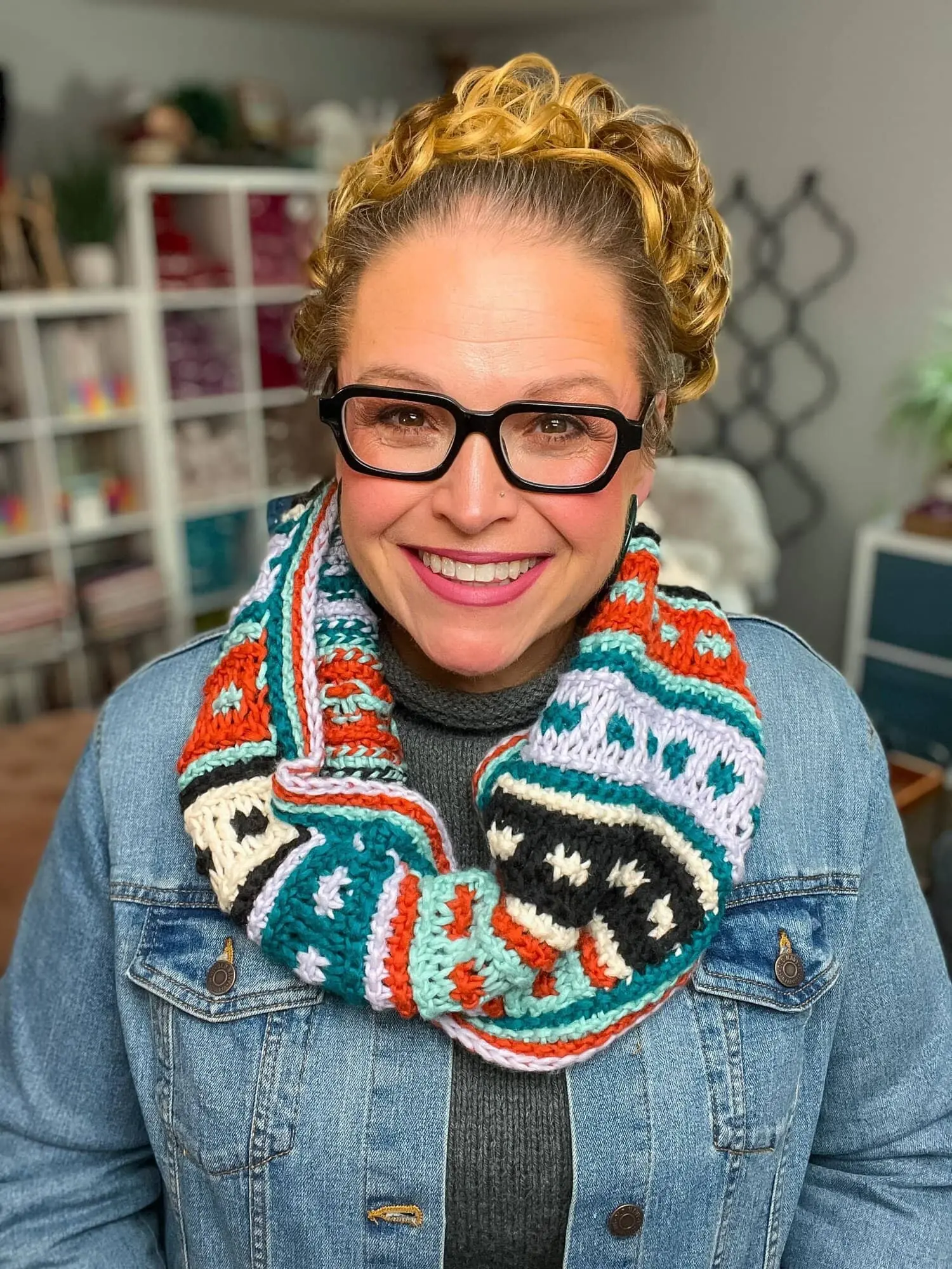 A smiling woman with glasses and curly blonde hair wears a colorful Tunisian crochet cowl pattern, a gray turtleneck, and a denim jacket. She stands indoors with shelves and yarn in the background, perfect inspiration for your next free pattern. -Marly Bird