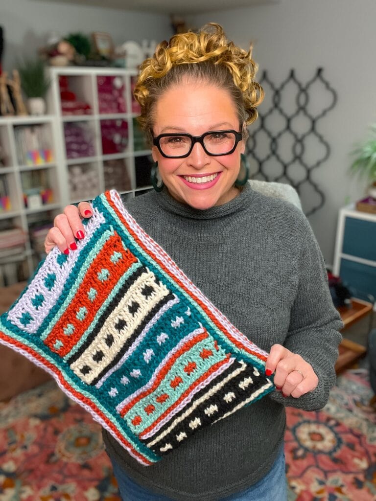 A smiling person with glasses and curly hair holds up a colorful Tunisian Crochet piece with multiple patterns and colors in a cozy room, shelves, and yarn in the background. -Marly Bird