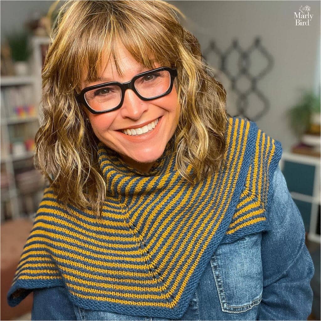 Blue and yellow striped knit shawl draped over a denim jacket, showing even stitches and soft texture. Shelves in background.