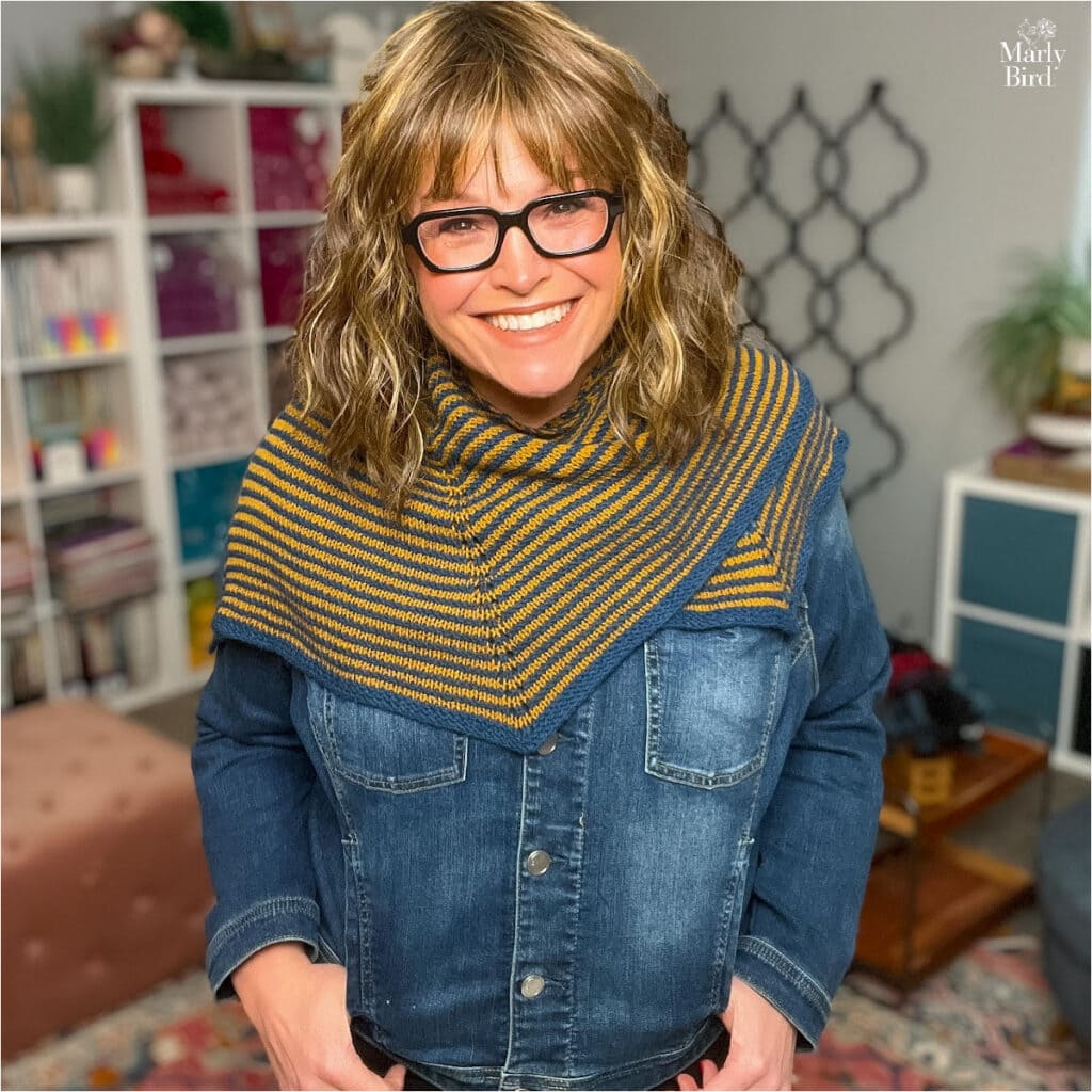 Blue and mustard striped knit shawl with textured stitches, draped over a denim jacket in a cozy craft room setting.