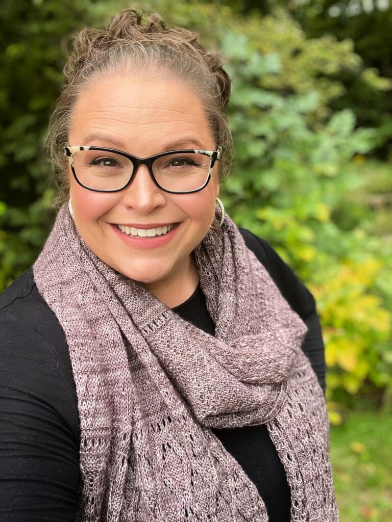 Woman models a knitted mosaic triangle shawl outdoors, showing off textured stitches against a lush green backdrop.