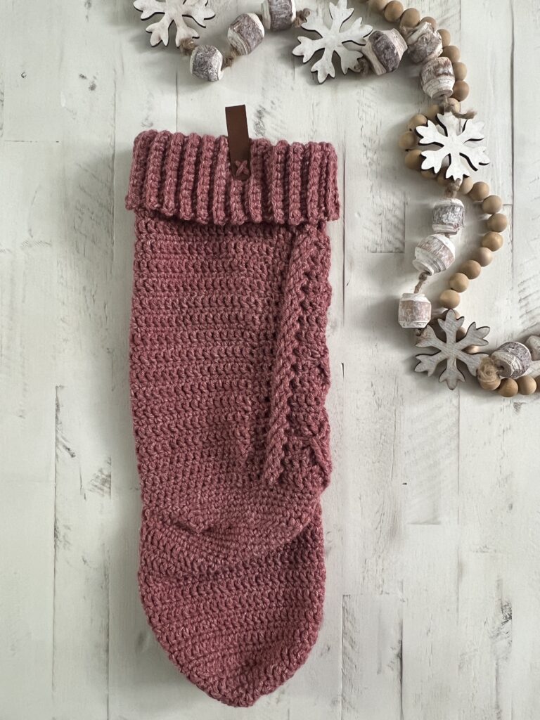 A knitted pink stocking with a folded cuff hangs on a white wooden surface, next to a decorative garland of wooden beads, snowflakes, and acorns—perfect inspiration for your next Crochet Christmas Stocking Pattern. -Marly Bird