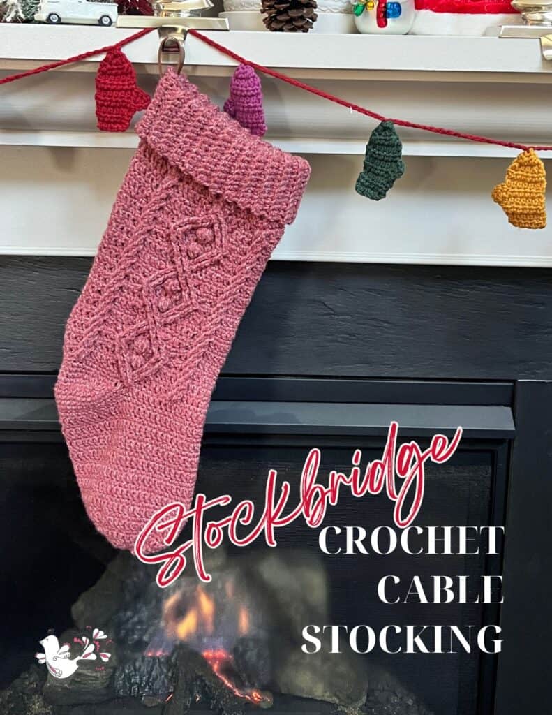 A pink crocheted stocking hangs from a mantel, adorned with a string of colorful lights for a jolly holiday feel. Text reads "Stockbridge Crochet Cable Stocking." A small dove graphic is at the bottom, reflecting Marly Crocheting's 2024 collection. -Marly Bird