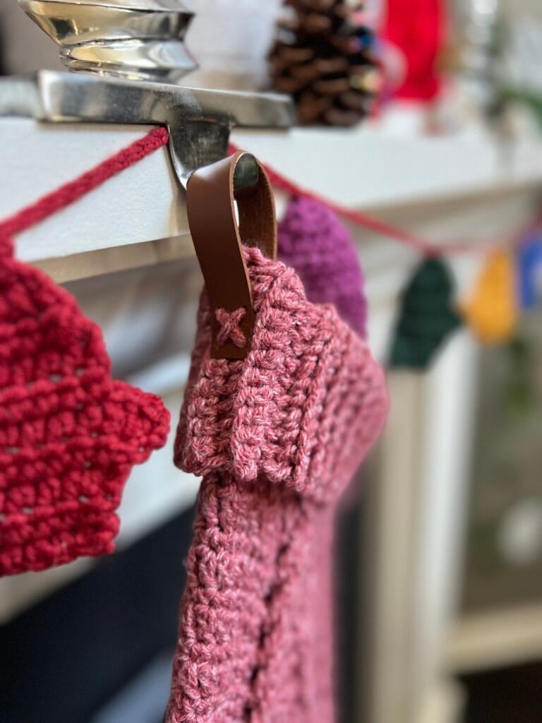 A pink knitted Christmas stocking with a brown leather loop hangs from a mantel. Red and green crocheted garland and festive decorations are visible in the background, perfect inspiration for your next Crochet Christmas Stocking Pattern. -Marly Bird