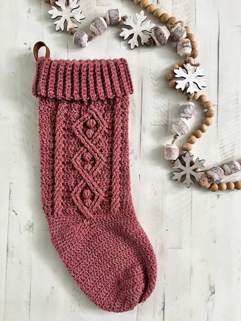 A knitted pink Christmas stocking with a diamond pattern, inspired by a Crochet Christmas Stocking Pattern, hangs beside a garland of wooden beads and white snowflake ornaments on a white wooden surface. -Marly Bird