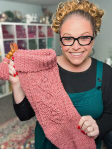 Woman displays a large pink knitted Christmas stocking with a textured stitch pattern; festive decor in background.