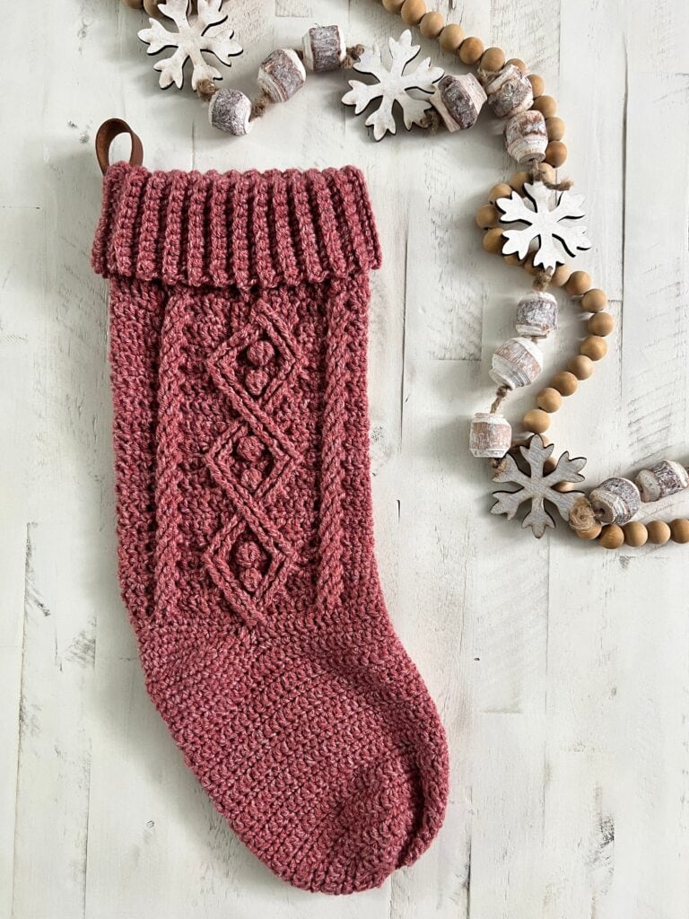 A knitted pink Christmas stocking with a folded cuff hangs on a white wooden background, decorated with a garland of wooden beads and white snowflake ornaments—perfect inspiration for those seeking free knitting and crochet patterns. -Marly Bird