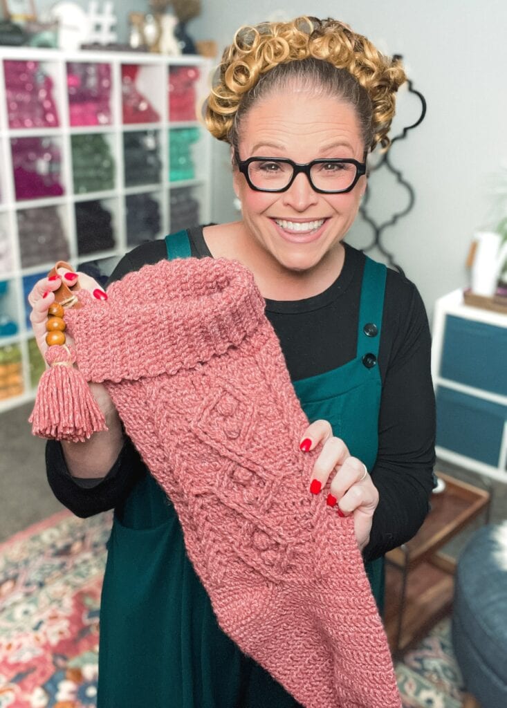 A smiling woman with glasses and curly hair holds up a large pink knitted stocking with a tassel in a cozy room filled with colorful yarn shelves, craft supplies, and inspiration for free knitting and crochet patterns. -Marly Bird