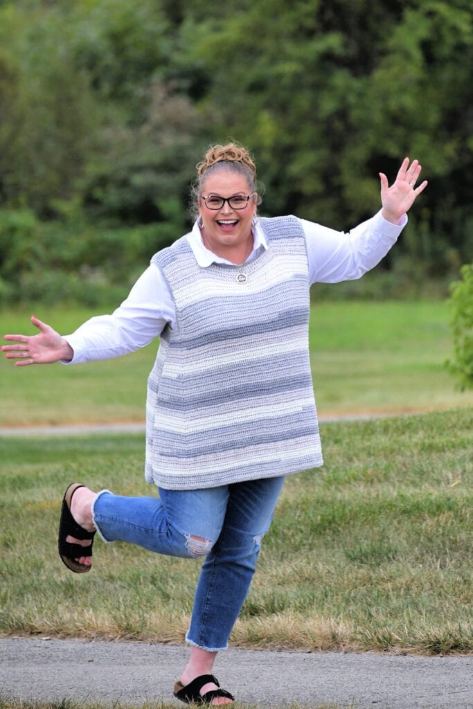 Marly Bird is wearing the Stoney Creek Sleeveless Crochet Tee over a white button down shirt and a pair of blue jean cropped jeans and birkenstock sandals. She has her hands out to her sides in jubilation and her right leg kicked up behind her like she is jumping for joy and excitement of this simple crochet pattern - Marly Bird