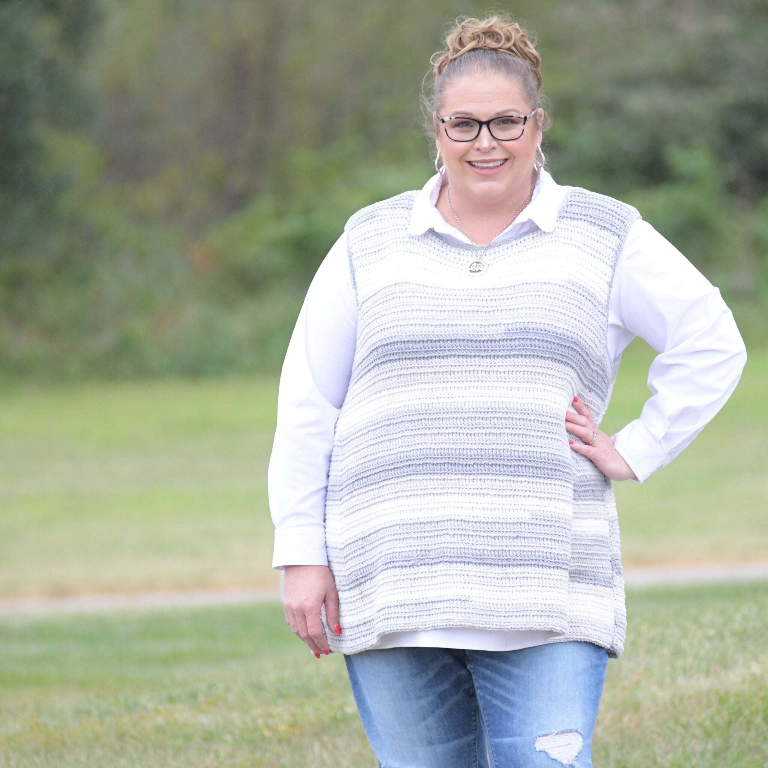 A woman models a gray-and-white striped crochet sweater, showing simple stitch texture and relaxed fit outdoors on grass.