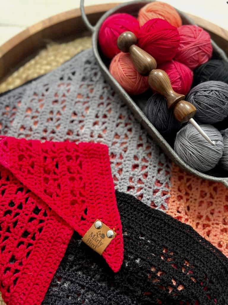 A set of colorful crochet yarn balls in a tray next to a folded, red crochet shawl with a leather tag. Other crocheted pieces in shades of grey, black, and light red are underneath. A wooden crochet hook rests on the yarn. -Marly Bird