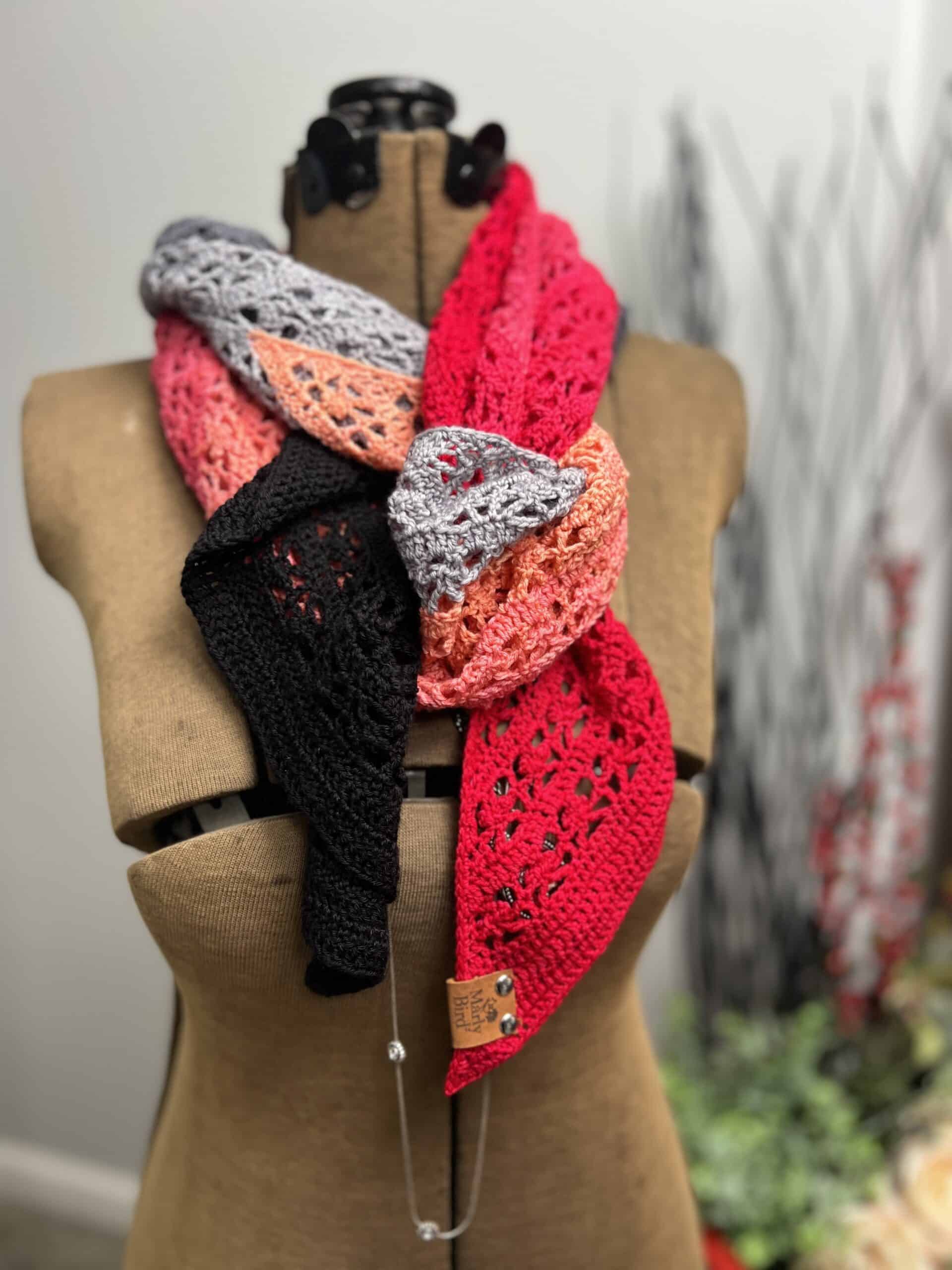 A beige mannequin displays a colorful, crocheted scarf with segments in red, pink, black, and gray patterns. The scarf is wrapped around the neck, showcasing its intricate design. Blurred background elements are visible. -Marly Bird