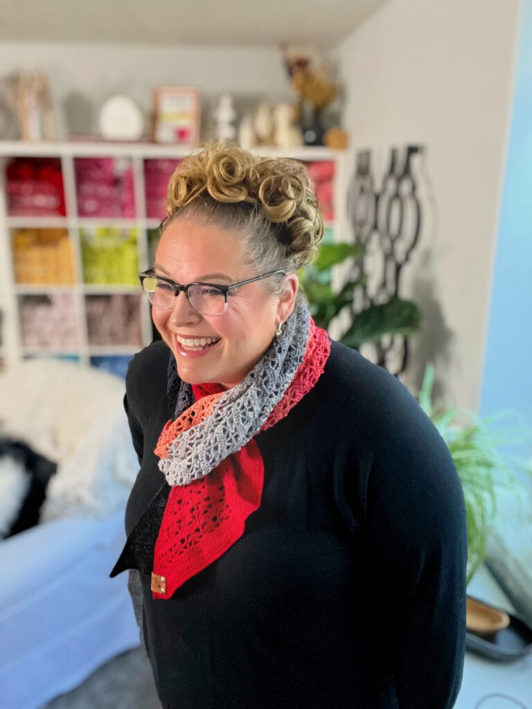 A person with curly hair and glasses smiles while wearing a multi-colored scarf and a black top. They stand in a cozy room with colorful shelves and plants. -Marly Bird